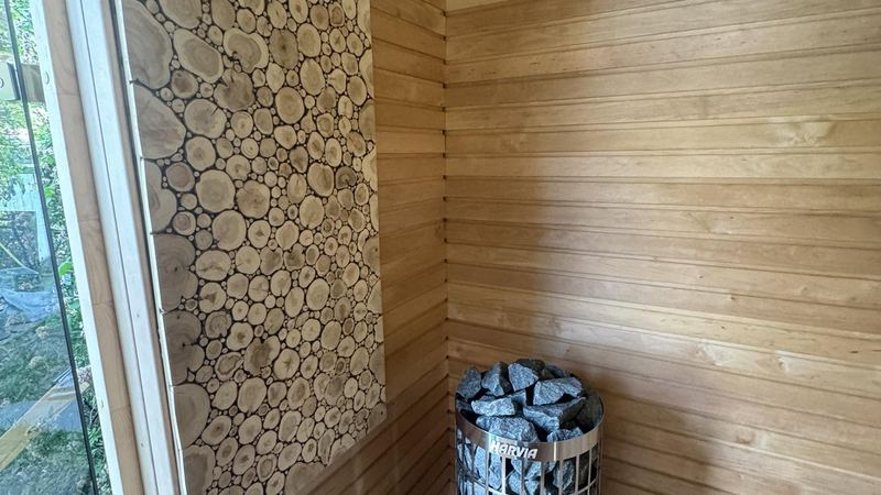 Cylindrical stone-filled sauna heater standing beside a decorative log-end wall panel