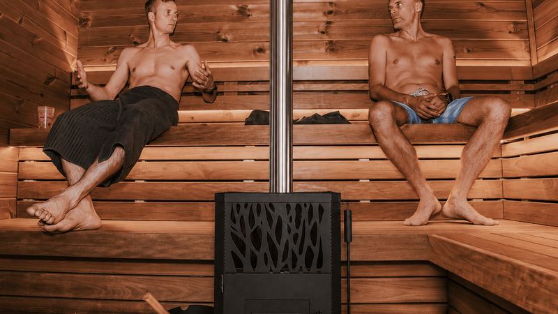 Two clients relaxing on opposite benches of a cedar sauna beside a wood-burning stove